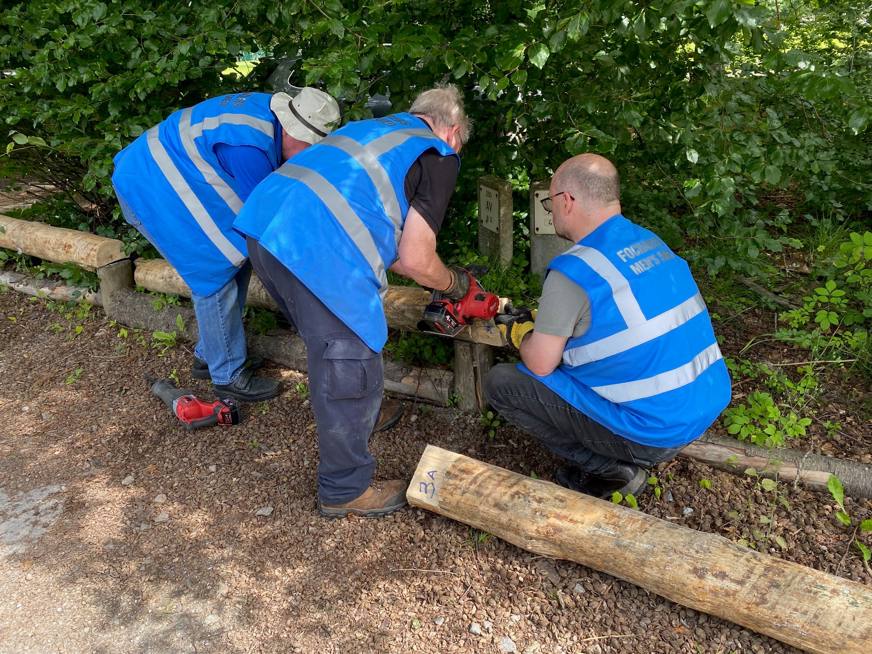Three men, wearing safety jackets with the wording Fochabers Men's Shed on the back, fix logs into place to create a woodland edging barrier. 