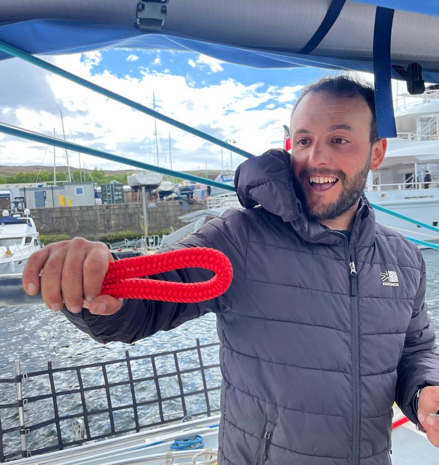 A person standing on the deck of a sailboat smiles while holding out a bright red braided rope with a spliced eye loop. They are wearing a dark insulated jacket, with marina docks, moored boats, and a cloudy blue sky visible in the background.