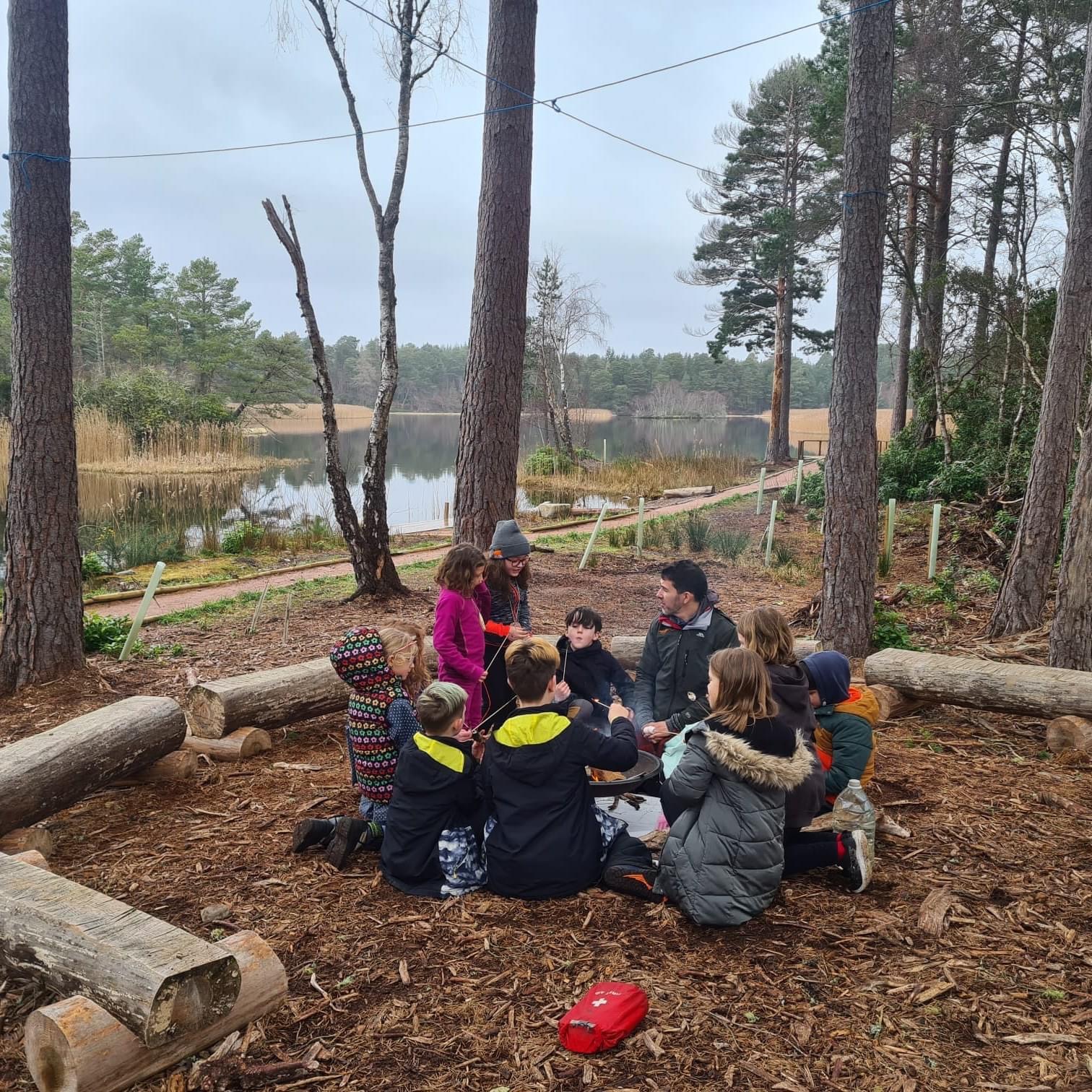 A group gathers around a fire, eating marshmallows, with Blairs Loch in the distance. 