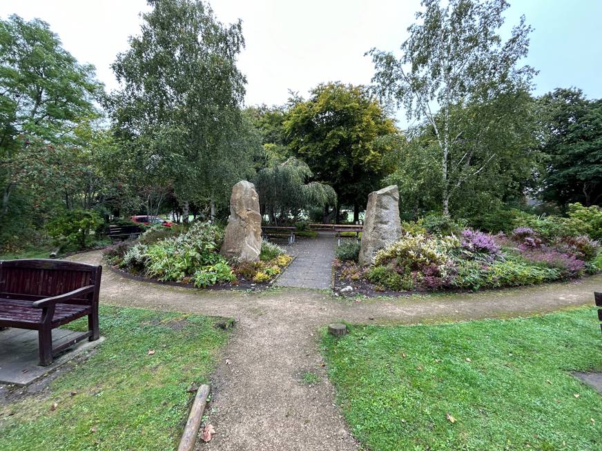 A garden pathway with tall stone monuments surrounded by lush greenery and vibrant flowers, complemented by wooden benches.