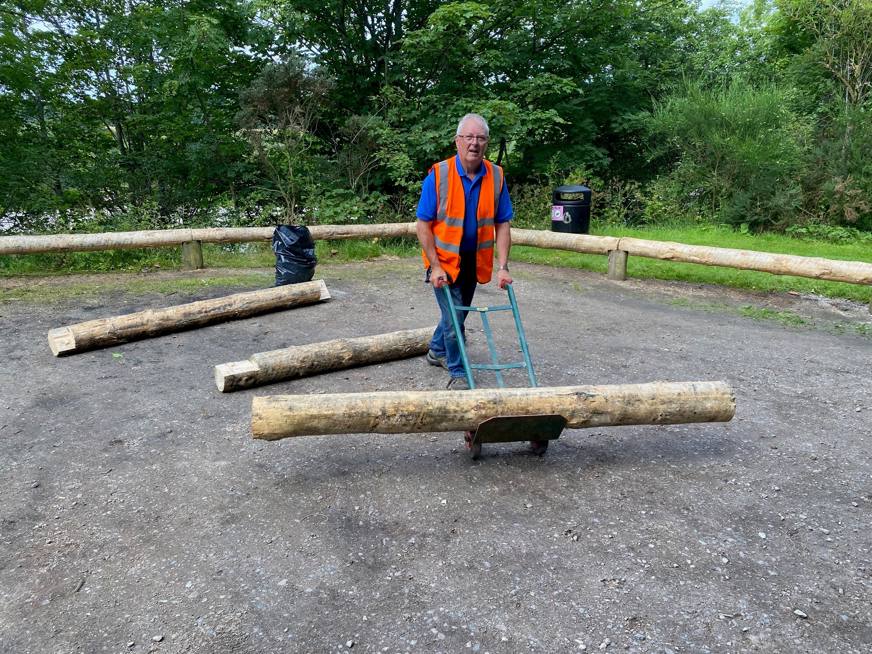 A man wearing a high visibility jacket pushes a trolley on which there is a large, shaped tree trunk. 