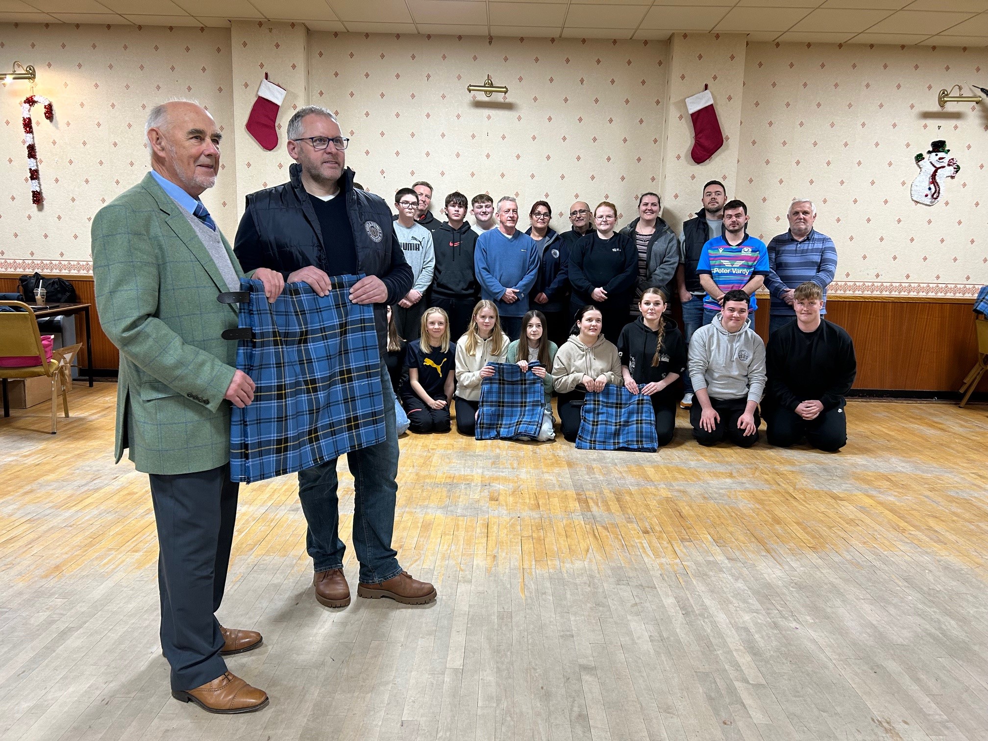 In a decorated indoor hall, two men stand in the foreground, holding a blue tartan kilt. Behind them, a group of people is gathered, smiling for the photo and also holding kilts.