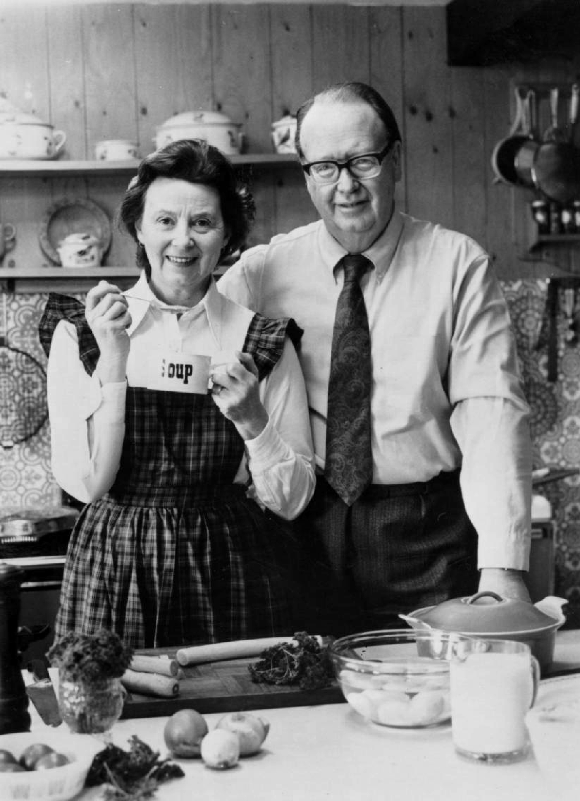 A black and white image featuring a couple, wearing tartan, in a kitchen where they are tasting soup. They smile as they look to the camera. 