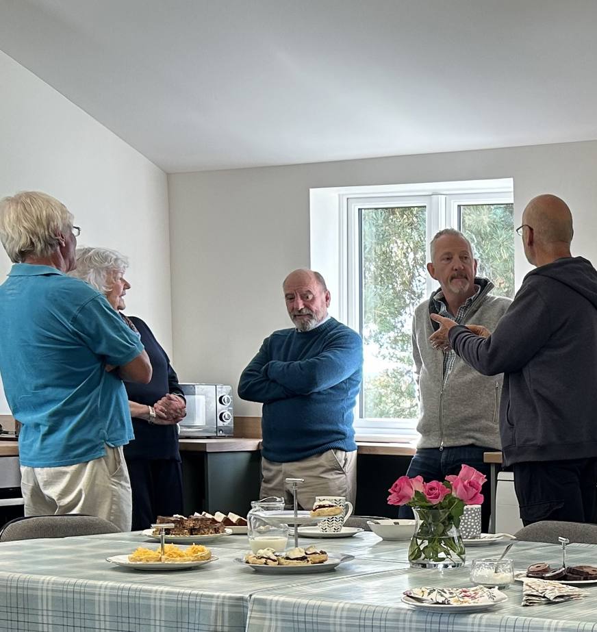 Pink roses sit in a glass vase in the middle of a large table, covered in a blue and white cloth and set for morning teas. There are cake, biscuits and scones. In the background a group of men and woman stand talking to each other. 