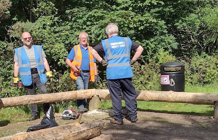 The sun shines as three members of Fochabers Men's Shed view their work. Beside them is an edging barrier, made from large planks of wood. 