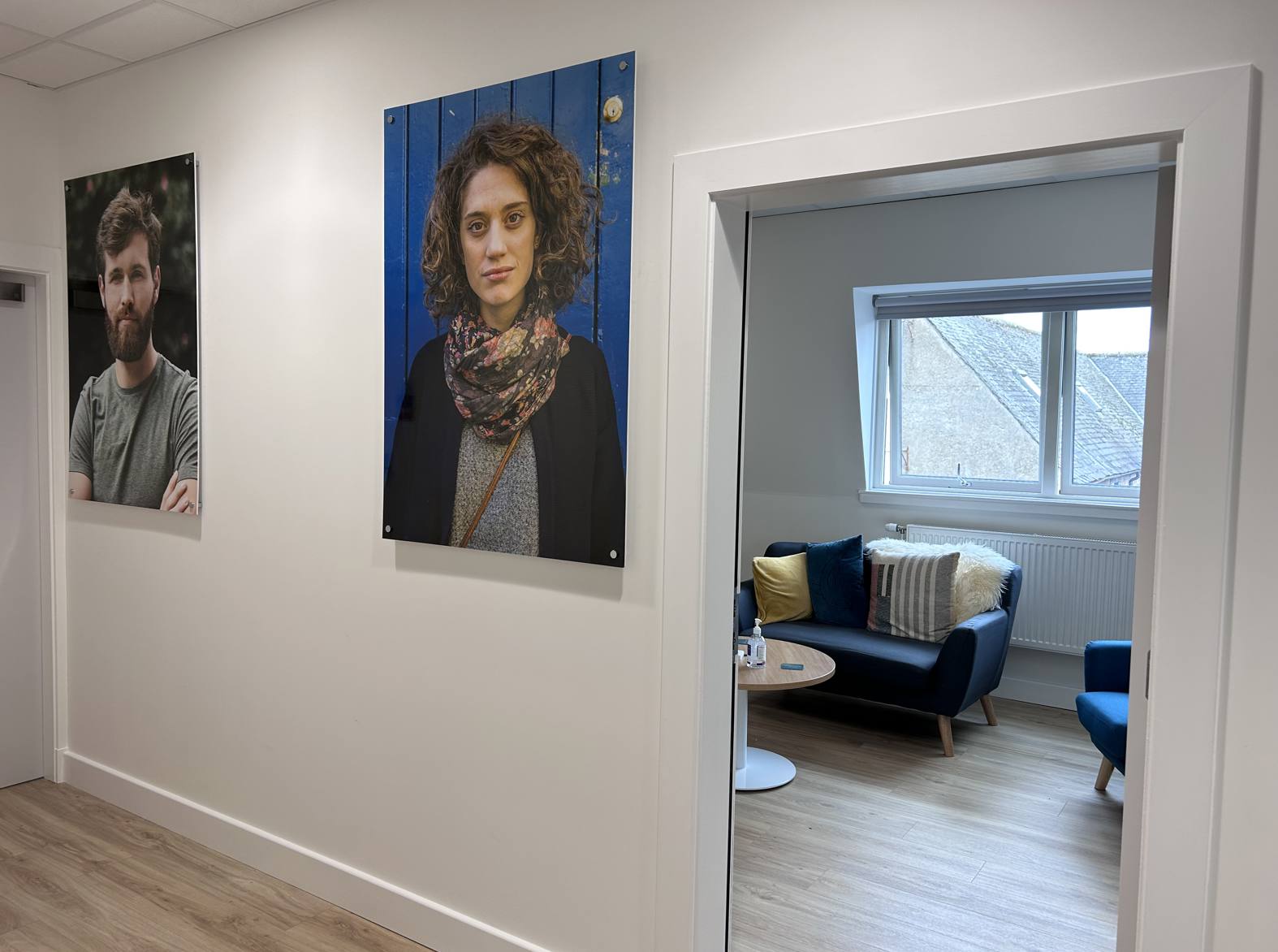 A bright, modern corridor leading to a welcoming support room. Two large portrait photographs hang on a white wall, while an open doorway reveals a comfortable meeting space with blue armchairs, a sofa, a round coffee table, wood-effect flooring, and a large window providing natural light. The space has a calm, professional atmosphere suitable for private meetings and support sessions.