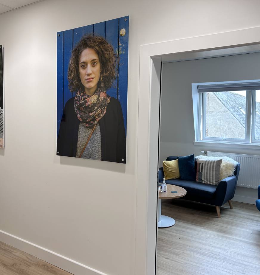 A bright, modern corridor leading to a welcoming support room. Two large portrait photographs hang on a white wall, while an open doorway reveals a comfortable meeting space with blue armchairs, a sofa, a round coffee table, wood-effect flooring, and a large window providing natural light. The space has a calm, professional atmosphere suitable for private meetings and support sessions.